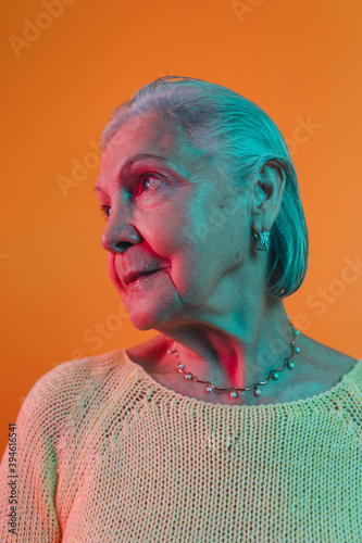 Thoughtful nice old woman with earings and pearls necklace looks away from camera isolated over orange studio background. Three quaters angle view. Peaceful old age concept. photo