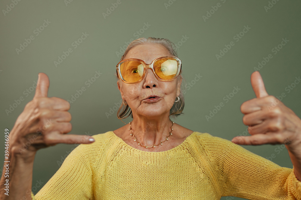 Very emotional elderly lady showing shaka sign and looking at camera. Skinny  pop eyed old woman in casual yellow shirt posing isolated over green  blackground. Orange glasses. Gray short hair. Stock 写真