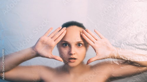 Face contouring. Defocused female portrait. Skin rejuvenation. Wrinkled texture scared woman with nude makeup bare shoulders looking through transparent film isolated on light background out of focus.
