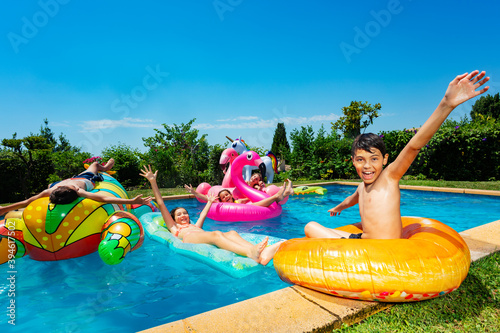 Many friends, boy in group of children play and have fun in swimming pool outside pose with inflatable doughnut toy ring lift hands