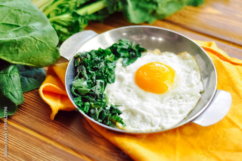 Green cooked spinach with fried egg on iron pan plate. Fried eggs healthy breakfast on wooden table background on orange napkin with spinach leaves, ingredients. Close up