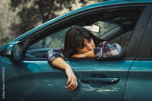 close-up of young woman with light skin and brown hair inside a car leaning against the window lowered of the car looking back with one arm hanging