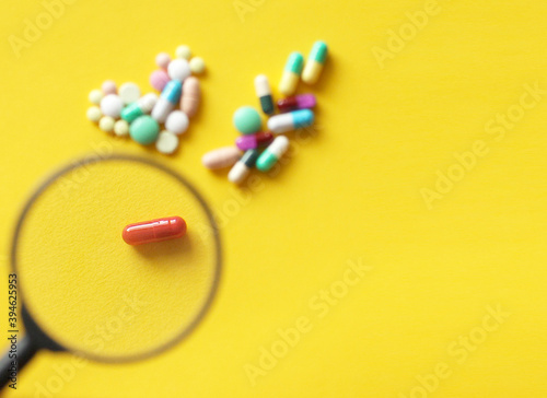 multicolored pills are scattered on a yellow background and a red pill is in a focus under a magnifying glass