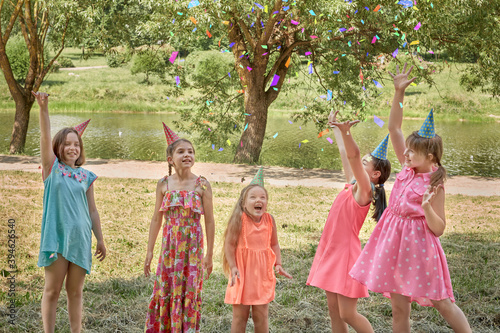 Girls throw up confetti at a children's party in a summer Park. Children in party hats. Large beautiful lake with reflection of trees.