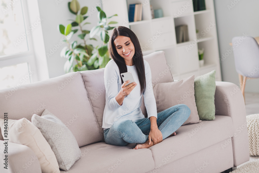 Full size photo of happy charming cheerful young woman wear white sweater jeans sit couch use phone inside home indoors Full size photo of happy charming cheerful young woman wear white sweater jeans sit couch use phone inside home indoors