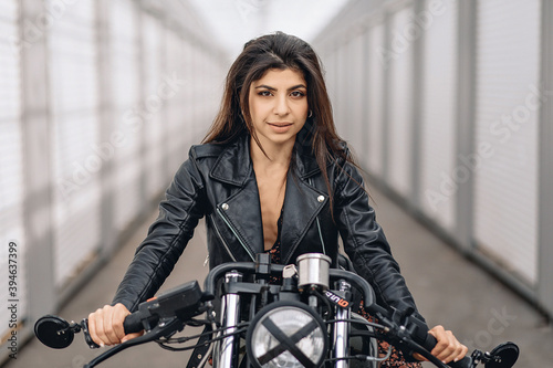 Portrait of a cheeky bright dark-haired model in a black leather jacket sitting on a black retro motorcycle and looking straight into the camera against a white background. Beauty concept