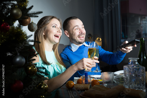 Cheerful young man and woman enjoying festive dinner and watching tv at Christmas night