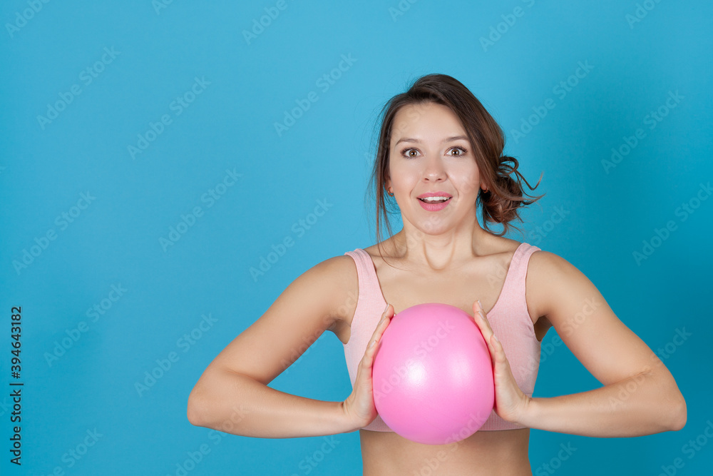 close-up portrait of a surprised girl in a pink sports top clutching a Pilates ball at her chest, isolated on a blue background. close-up portrait of a surprised girl in a pink sports top clutching a Pilates ball at her chest, isolated on a blue background.