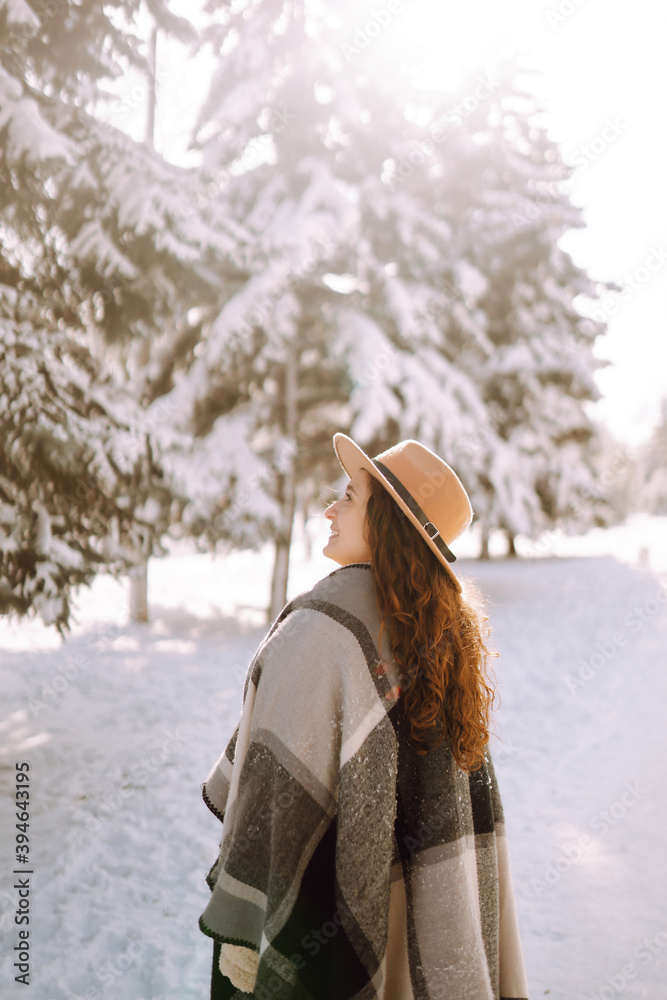 Fototapeta premium Beautiful woman in hat, plaid scarf and coat posing with joy outside in the snow forest. Cheerful curly lady enjoying winter moments in a snowy park. Holidays, season and leisure concept. Christmas.