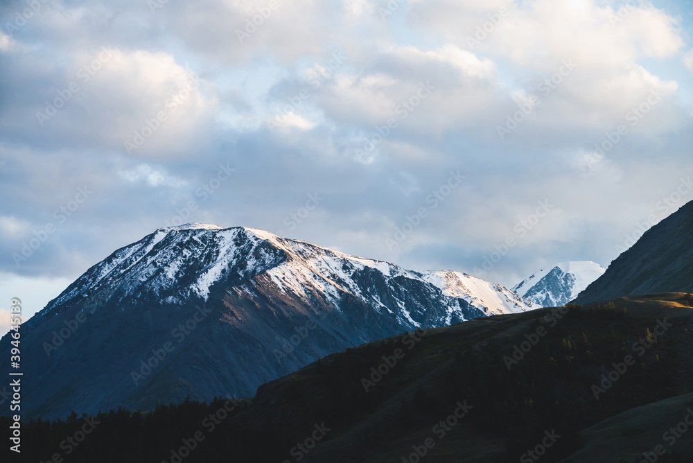 Atmospheric alpine landscape to snowy mountain ridge and forest hills in sunset. Snow shines in golden light on mountain peak. Wonderful scenery with beautiful shiny snowy top. Evening cloudy sky.