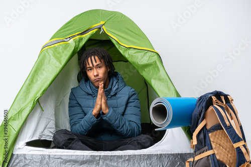 Young african american man inside a camping green tent keeps palm together. Person asks for something