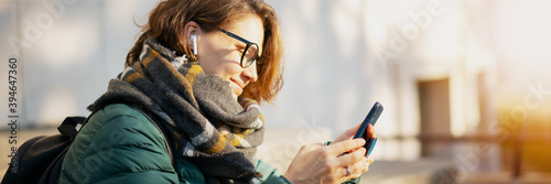 Young woman with wireless headphones and mobile device listening to music outdoor in autumn winter city