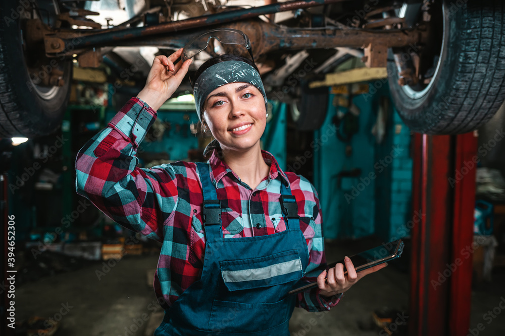 Portrait of a young happy female mechanic in uniform posing with tablet at her hands and remove glasses from the head. The car is located on the lift