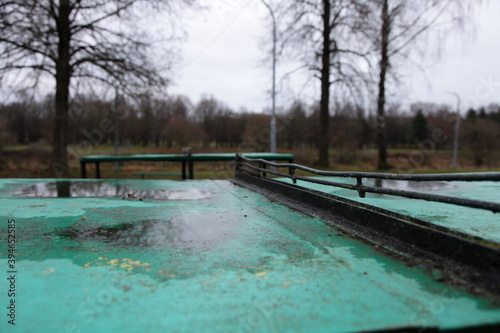 Empty old table tennis metal table with puddles in an abandoned sports field in the Park on a cloudy autumn day on bare trees background