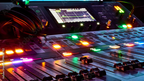 Low angle close up on a dark studio mixing sound board, with rows of small dials illuminated by a rainbow spot light, in the control booth at a concert. color