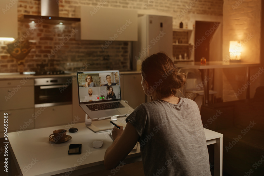 Back view of a female employee who is working remotely talking to her colleagues in a video conference on a laptop from home. A multiethnic business team on an online meeting in the evening. Back view of a female employee who is working remotely talking to her colleagues in a video conference on a laptop from home. A multiethnic business team on an online meeting in the evening.