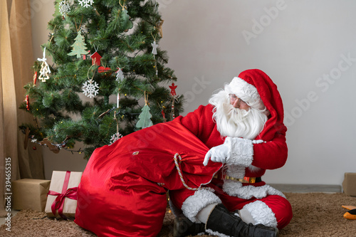 santa claus sits on the floor near the christmas tree unties the bag and takes out a gift