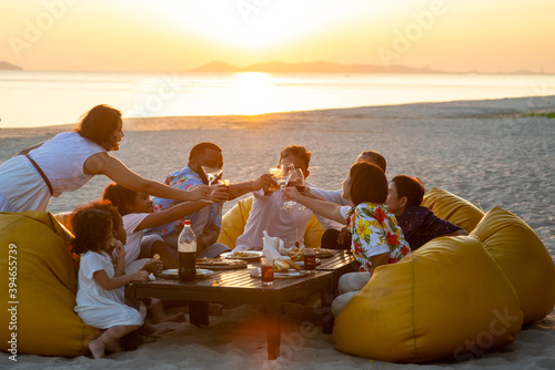 Group of multiethnic family friends enjoy dinner party together on the beach at sunset. Diverse family with child girl, adult and senior couple relax and having fun together on summer holiday vacation