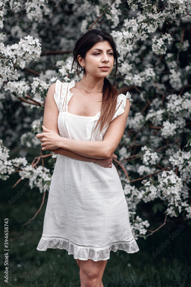 pensive brunette girl in a whire tight dress is posing near the tree of lilac, portrait pensive brunette girl in a whire tight dress is posing near the tree of lilac, portrait