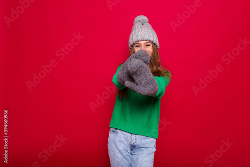Happy excited young woman with long hair wearing knitted sweater and cap covering face with hands over red background