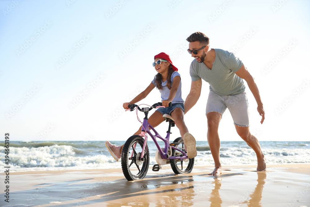 Obraz premium Happy father teaching daughter to ride bicycle on sandy beach near sea