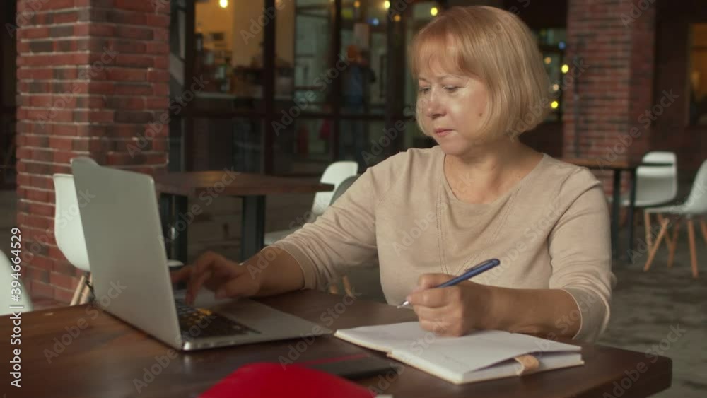 Senior woman works on laptop and writes down something in notebook at the modern cafe Senior woman works on laptop and writes down something in notebook at the modern cafe