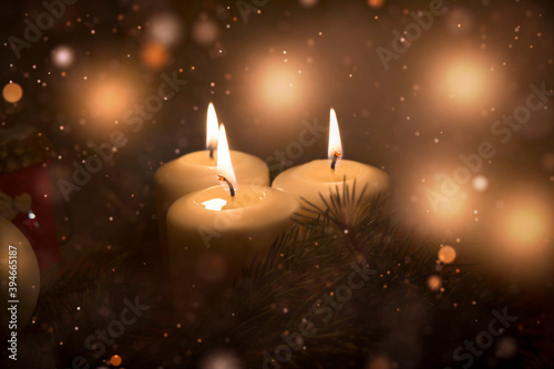 White Christmas candles are burning, green fir branches are lying next to them, Christmas toys, festive highlights of garlands and the flame of three candles give a sense of celebration.
