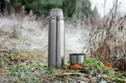 Vacuum thermos with a mug in the winter forest. Steel travel flask and a glass of steam. Concept for hot winter drink, travel, hike.