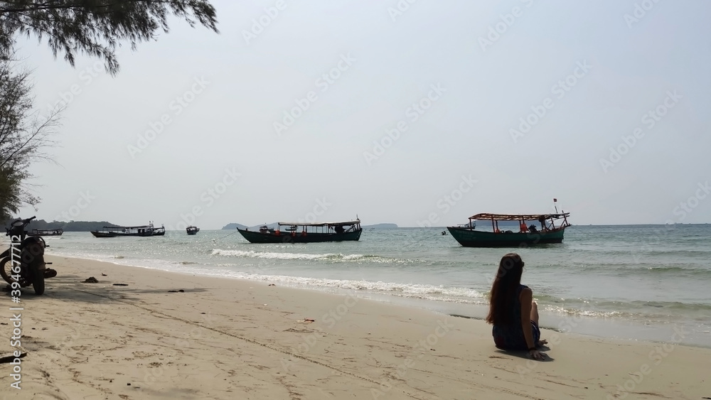 The girl is sitting on the beach. Motorcycle and fishing boats at sea. Otres 2. Sihanoukville. Cambodia. South-East Asia