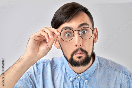 Wow! Caucasian bearded man with bulging eyes and a surprised face holds hands on the rim of glasses and looks at the camera, emotional reaction to unexpected news isolated in blue studio