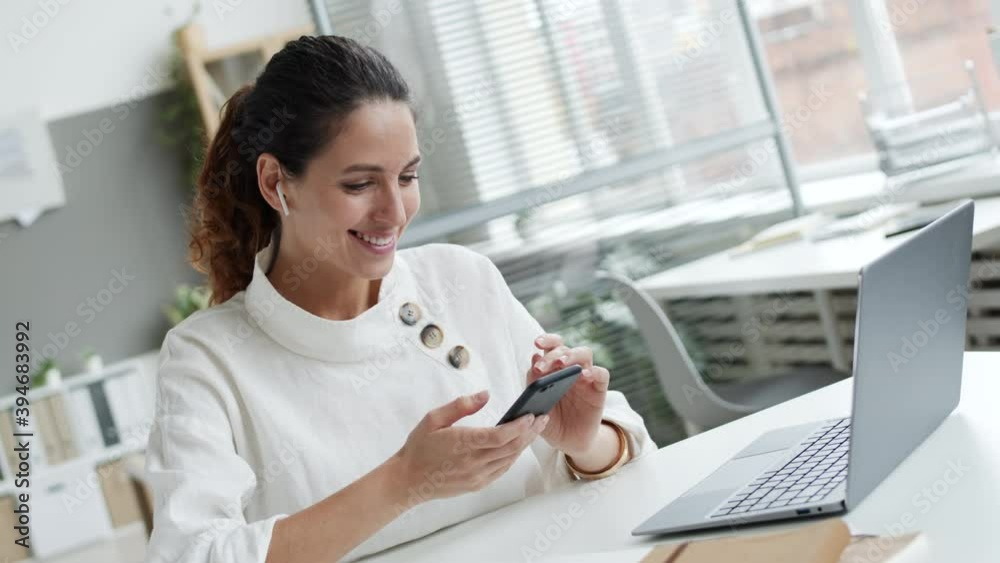 Medium shot of young Caucasian woman with curly hair wearing wireless headphones sitting at workplace in front of laptop and using smartphone Medium shot of young Caucasian woman with curly hair wearing wireless headphones sitting at workplace in front of laptop and using smartphone