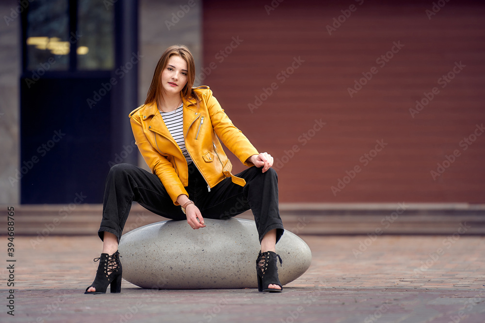 Fototapeta premium A young woman sits on a city cobblestone, a large stone. She is wearing high heels and a yellow jacket.