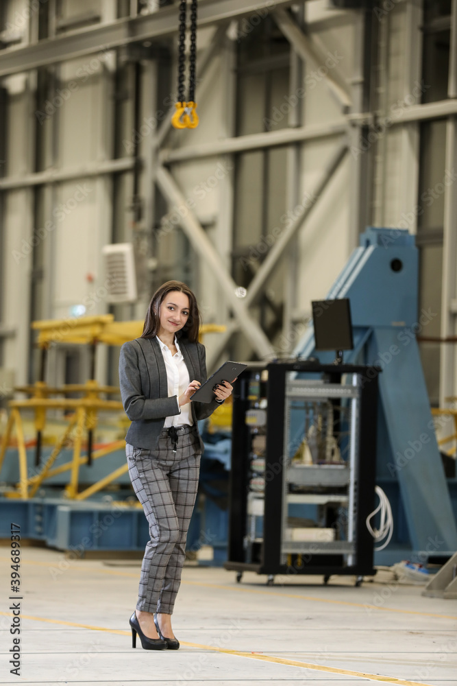 Portrait of a female factory manager in a white hard hat and business suit holding mobile phone and tablet , controlling the work process in the helicopter manufacturer.