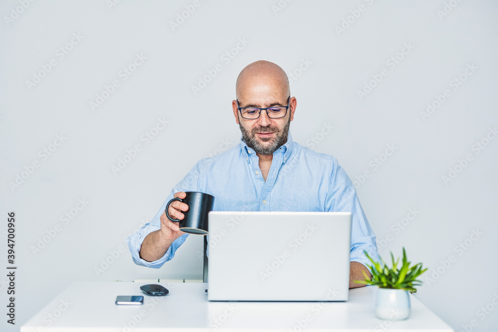 Businessman teleworking with a laptop having a coffee. Bald man with glasses