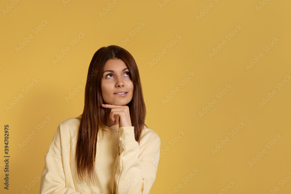 Thoughtful caucasian woman in yellow hoodie looks pensively upwards, has pleasant dreams planing new deals, isolated over a yellow background with copy space for your advertising. Thoughtful caucasian woman in yellow hoodie looks pensively upwards, has pleasant dreams planing new deals, isolated over a yellow background with copy space for your advertising.