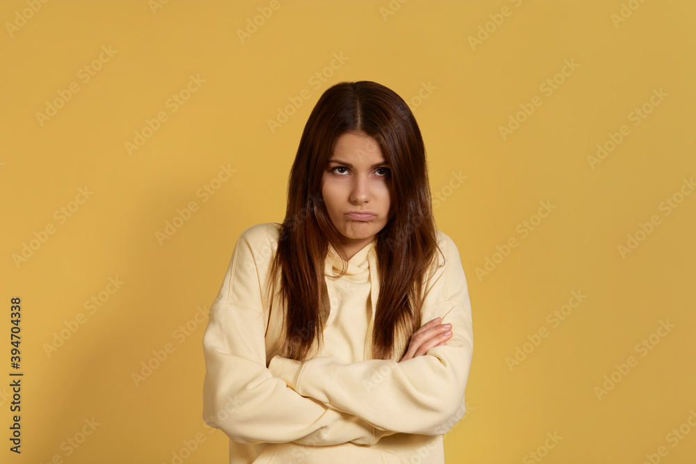 In furious. Sullen caucasian woman in yellow hoodie, keeps arms folded, has sullen expression, being discontent with results of something. Has troubles with boyfriend. Isolated on a yellow background. In furious. Sullen caucasian woman in yellow hoodie, keeps arms folded, has sullen expression, being discontent with results of something. Has troubles with boyfriend. Isolated on a yellow background.