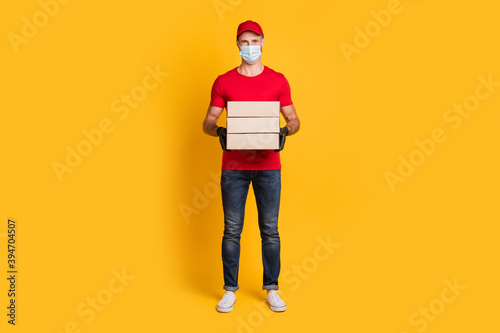 Photo of young courier guy hold pile boxes wear mask red t-shirt hat jeans footwear isolated yellow color background