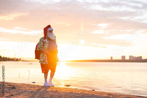 Fototapeta Naklejka Na Ścianę i Meble -  Santa claus in shorts and a shirt walks along the beach at sunset. Christmas in a hot country