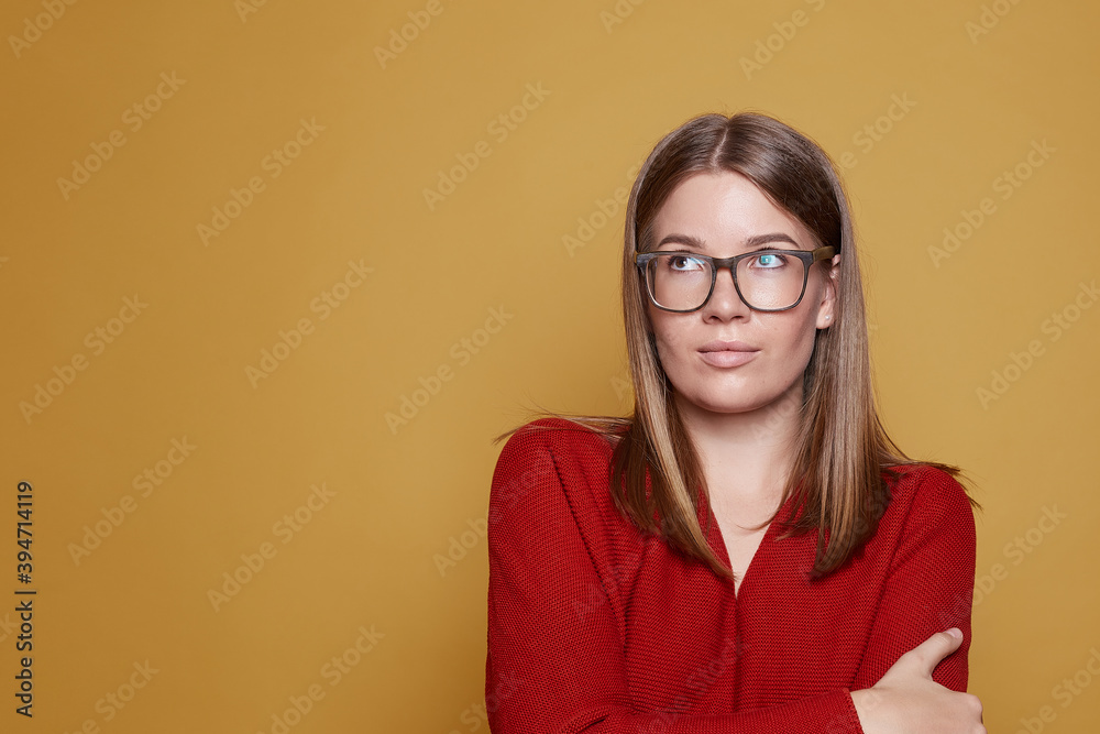 Shot of adorable self confident businesswoman or designer, stands crossed hands, looking up, wears casual red blouse and spectacles, poses against yellow studio wall with copy space. Lifestyle concept