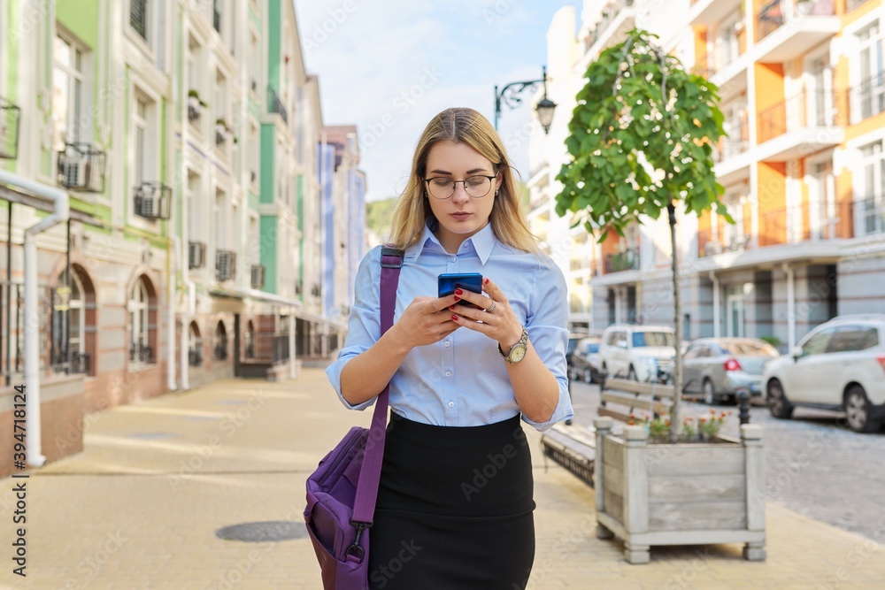 Fototapeta premium Young business woman with smartphone walking along city street