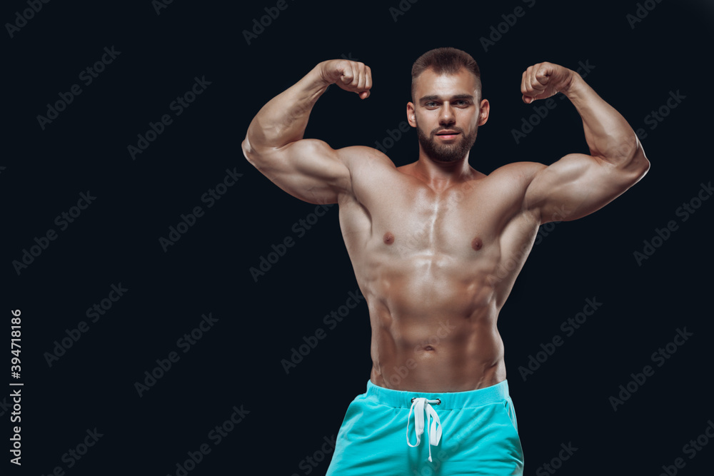 Fototapeta premium Front view of muscular and fit young bodybuilder fitness male model demonstrating his biceps isolated on black background