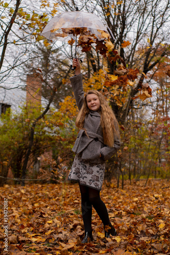 cute beautiful teenage brunette girl in an grey coat holding a transparent umbrella with falling leafes over her. Cosiness, autumn