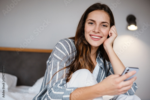 Pleased brunette woman using cellphone while sitting on bed