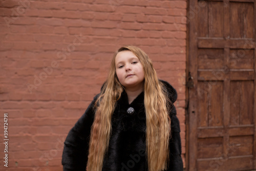 portrait of cute brunette teen girl in black fur coat on the background of brown brick wall ans old wooden door . Cosiness, fashion, style