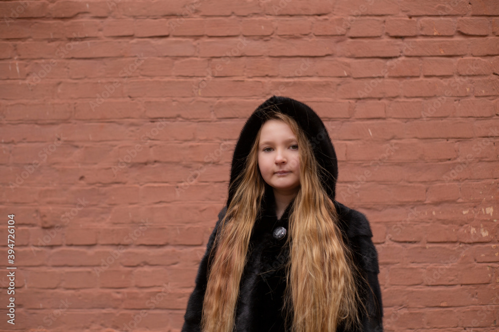 Naklejka premium portrait of cute brunette teen girl in black fur coat on the background of brown brick wall . Cosiness, fashion, style