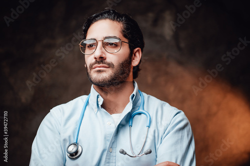 Serious doctor specialist with eyeglasses and dressed in white coat posing in dark background looking away.