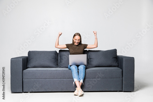 Happy young woman using laptop sitting at sofa with win gesture on white background