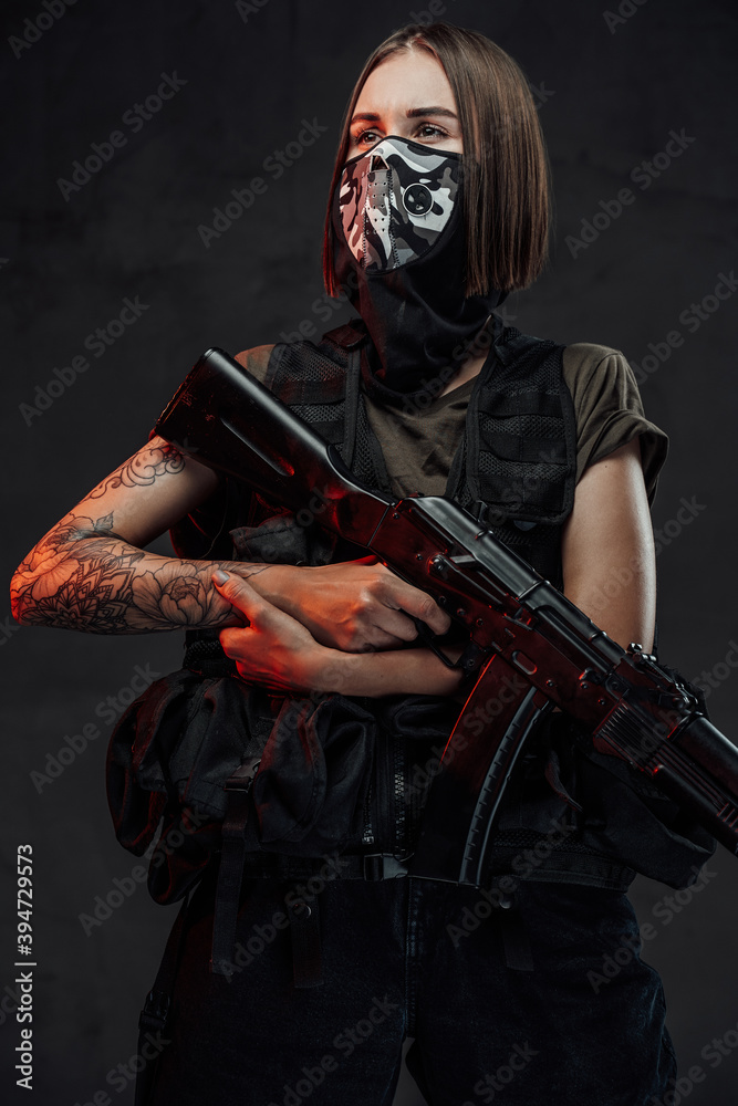 Armed with ak74 rifle and weared with mask stylish female mercenary poses in dark background.