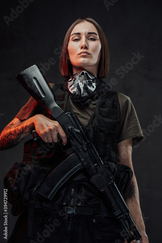 Stylish and martial woman with short haircut and tattooed arm poses holding ak74 rifle in dark background.