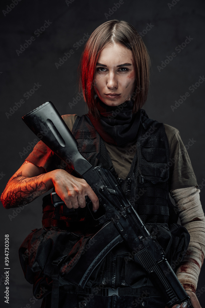 Dreseed in dark armour military woman with short haircut holds rifle with tattooed and bandaged hands in dark background. Dreseed in dark armour military woman with short haircut holds rifle with tattooed and bandaged hands in dark background.